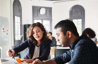 [Featured image] A male, wearing a dark shirt and a female, wearing a dark jacket, are working in front of a desktop as they discuss the change management process. 