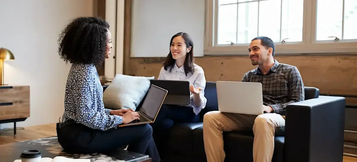 [Featured Image] Three business degree students sitting around a couch, looking at each other and smiling with open laptops in their laps.