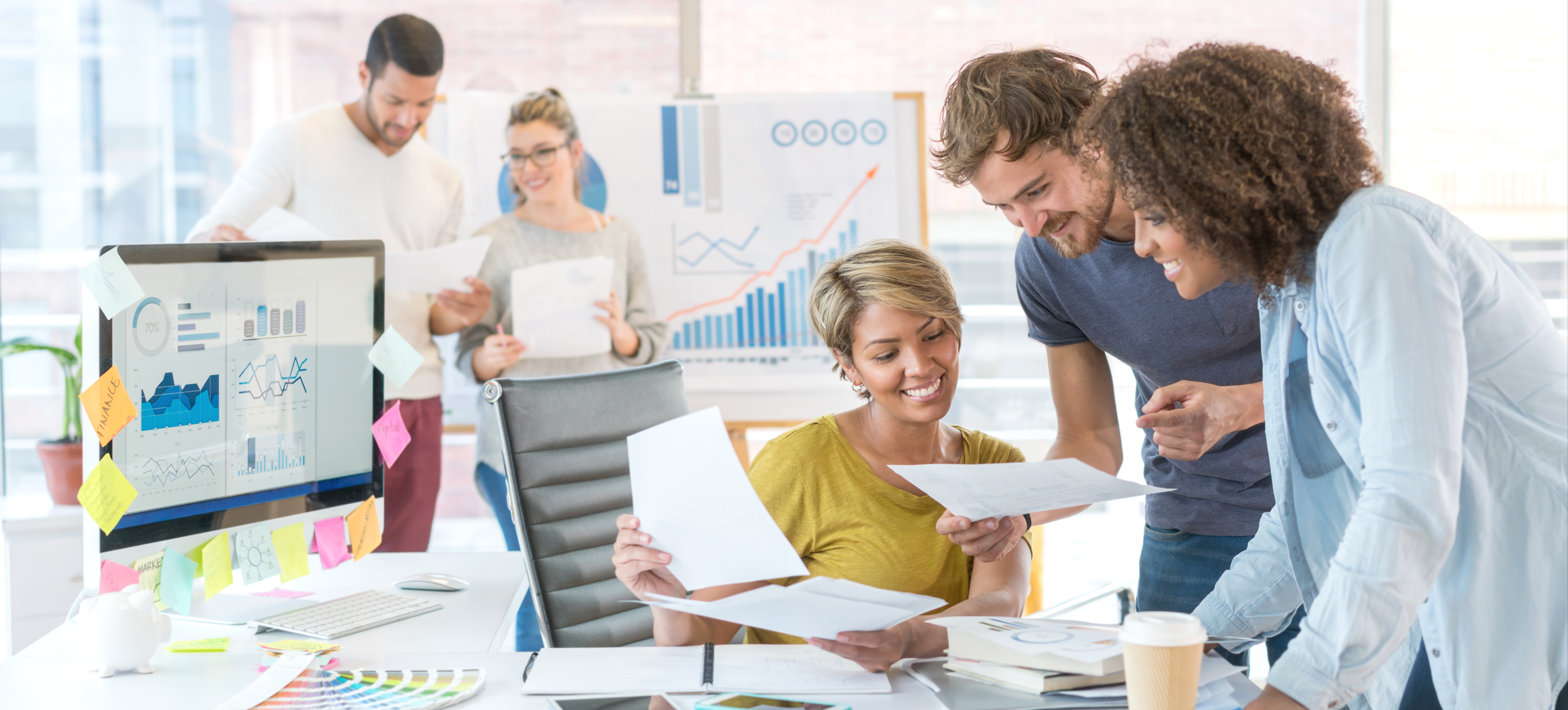 [Hauptbild] Eine glückliche Wachstumsvermarkterin und ihr Team besprechen ihre Wachstumsmarketingstrategie, während sie Daten auf Papier und einem Computerbildschirm durchsehen.