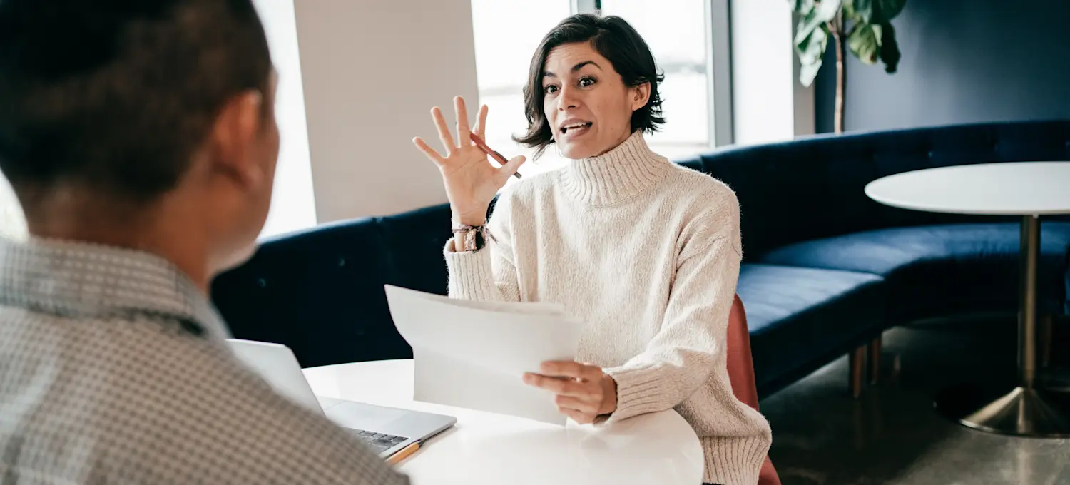 [Featured Image]: Two people sit facing each other at a round table. One person is holding a paper and asking email marketing interview questions to another person whose back is to the camera.

