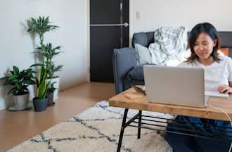 [Featured Image] A woman in a white t-shirt sits on the floor at her coffee table and types prompts into ChatGPT for a project.