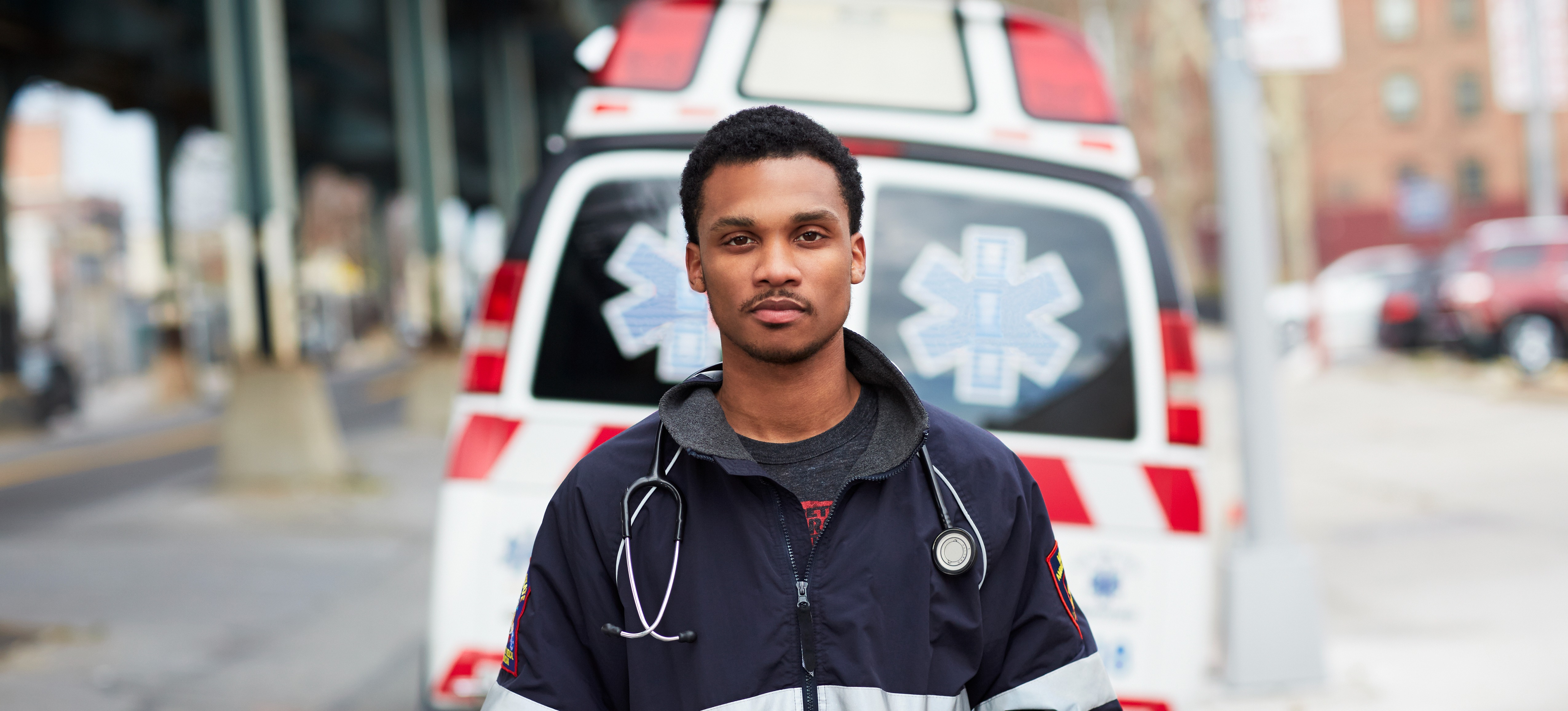 [Featured Image]: A person with short black hair is wearing a black and white jacket. They are standing in front of an ambulance. 
