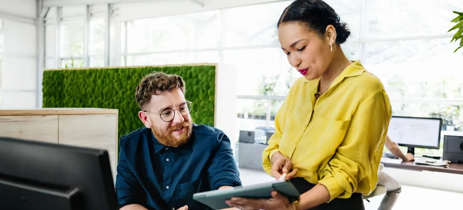 [Featured Image] An implementation manager holds a tablet out to show something to a seated colleague as they stands at their desk.