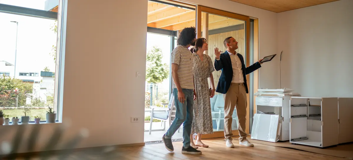 [Featured Image]: A real estate agent holding a realtor certification shows a property to a young couple planning to buy a home.
