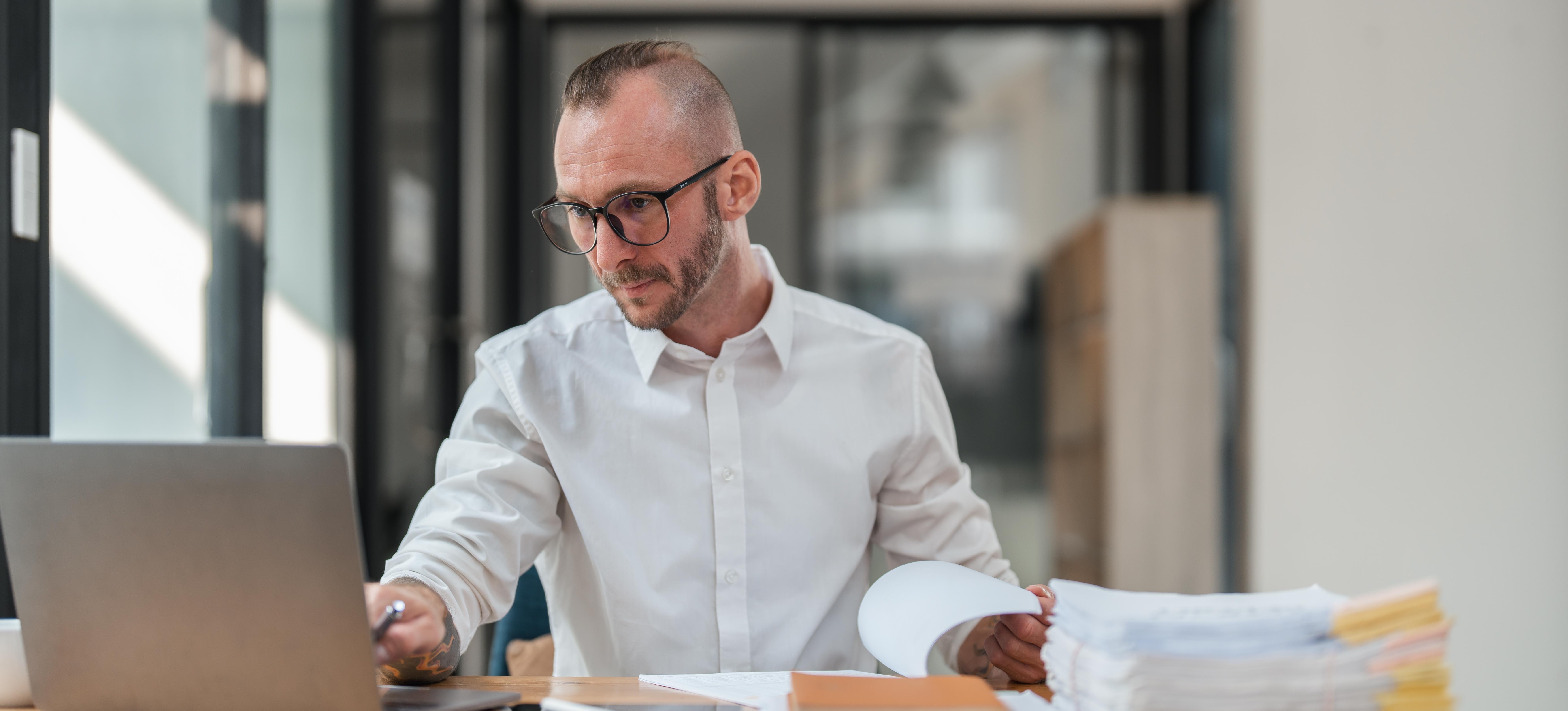 [Featured Image] A tax specialist compares paperwork to information on their computer with a stack of tax documents on their desk.