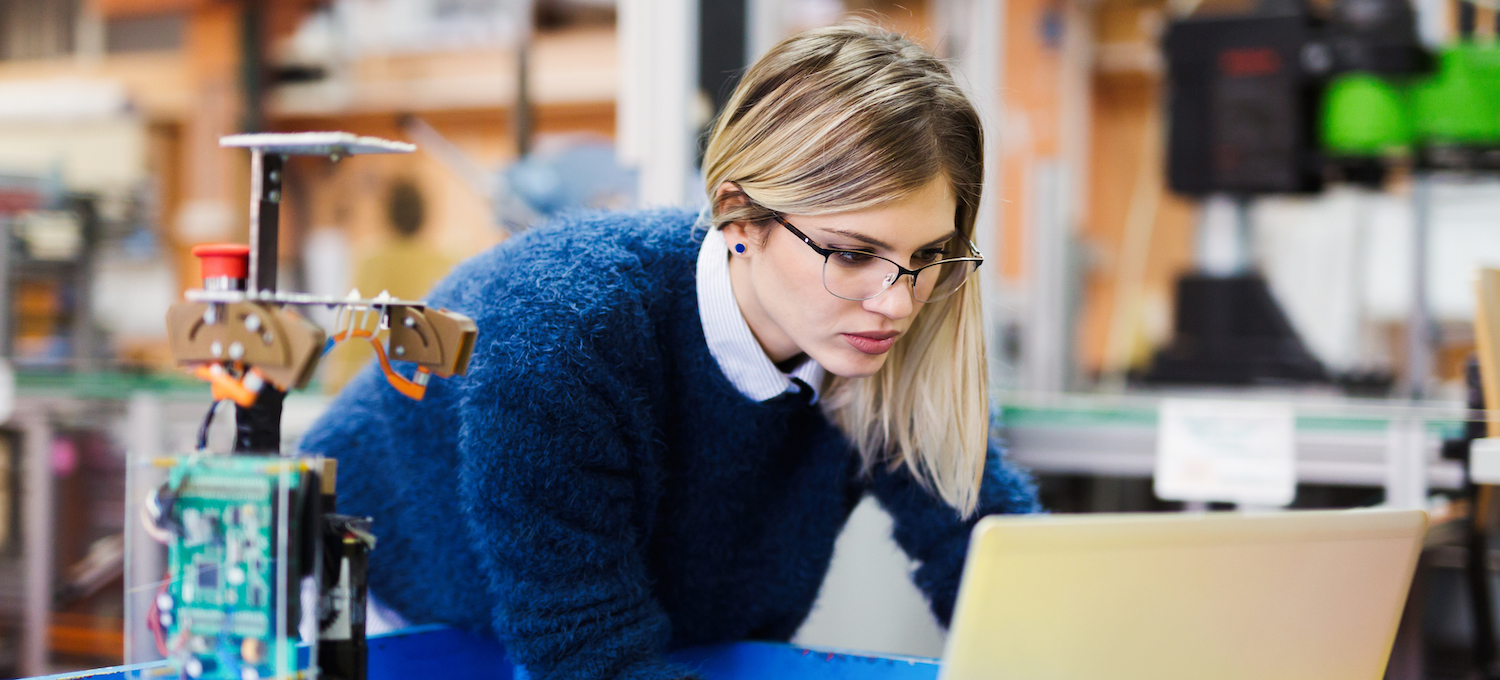 [Featured image] A student wearing glasses stands over a robotics work table staring at their laptop.