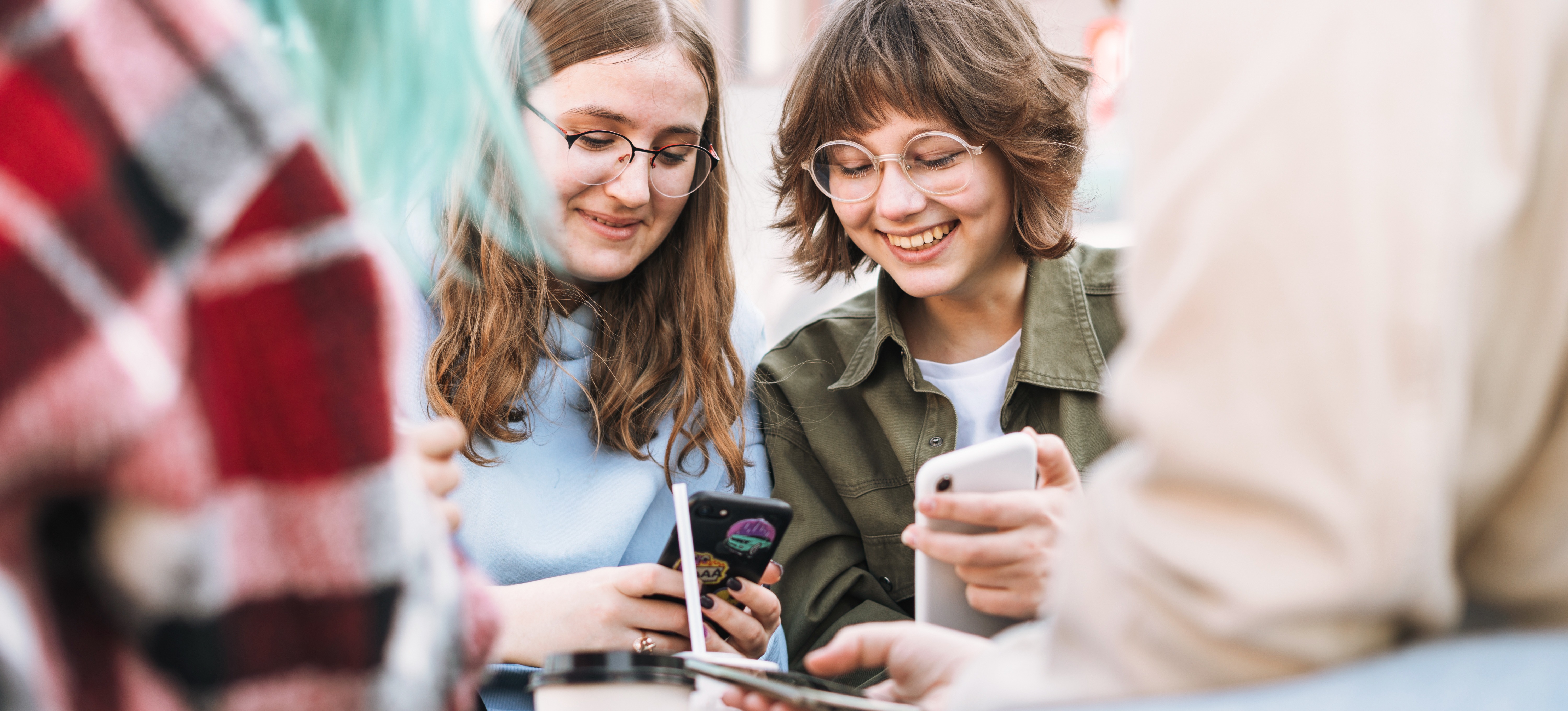 [Featured image] Two friends are at a table outside in the city looking at their phones and talking about going to college after earning their GED.