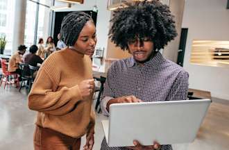 [Featured Image] Two coworkers review information on a laptop in an office.