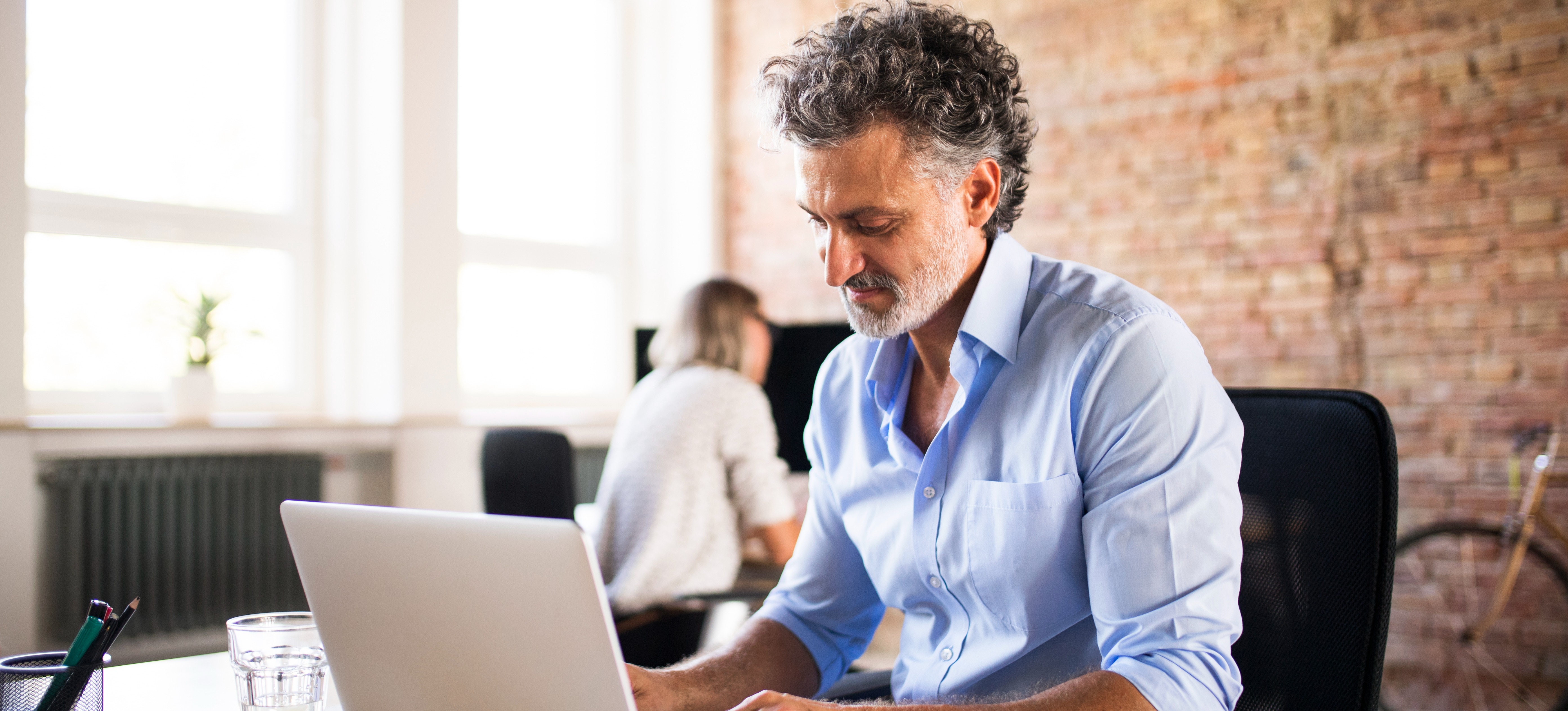 [Feature Image] An IT professional researches, “Which CompTIA exam should I take?” on their laptop computer in a room with a brick wall and ample natural light.