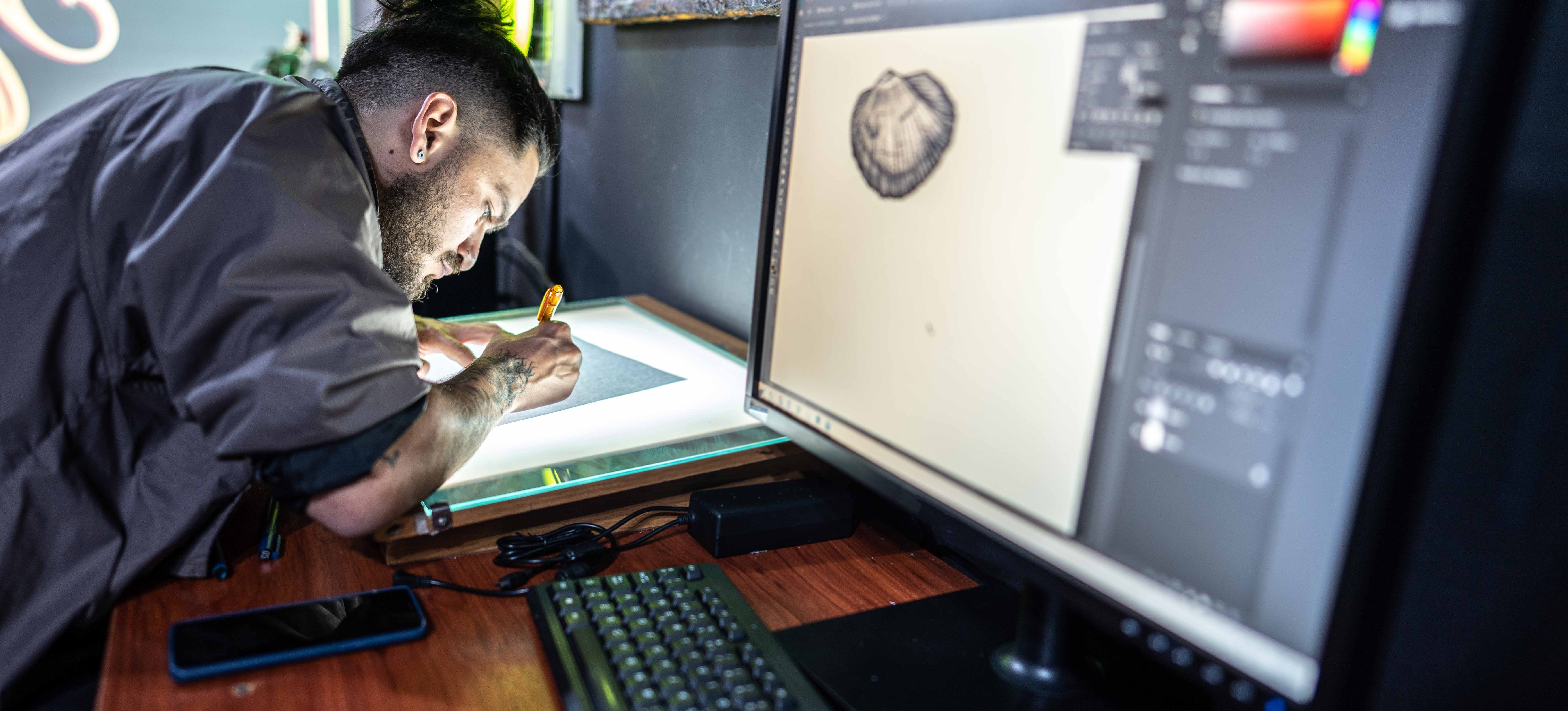 [Featured Image] An artist draws on paper in a tattoo studio with the image on a graphics designing application—showcasing creativity and tools that influence their graphic designer salary.
