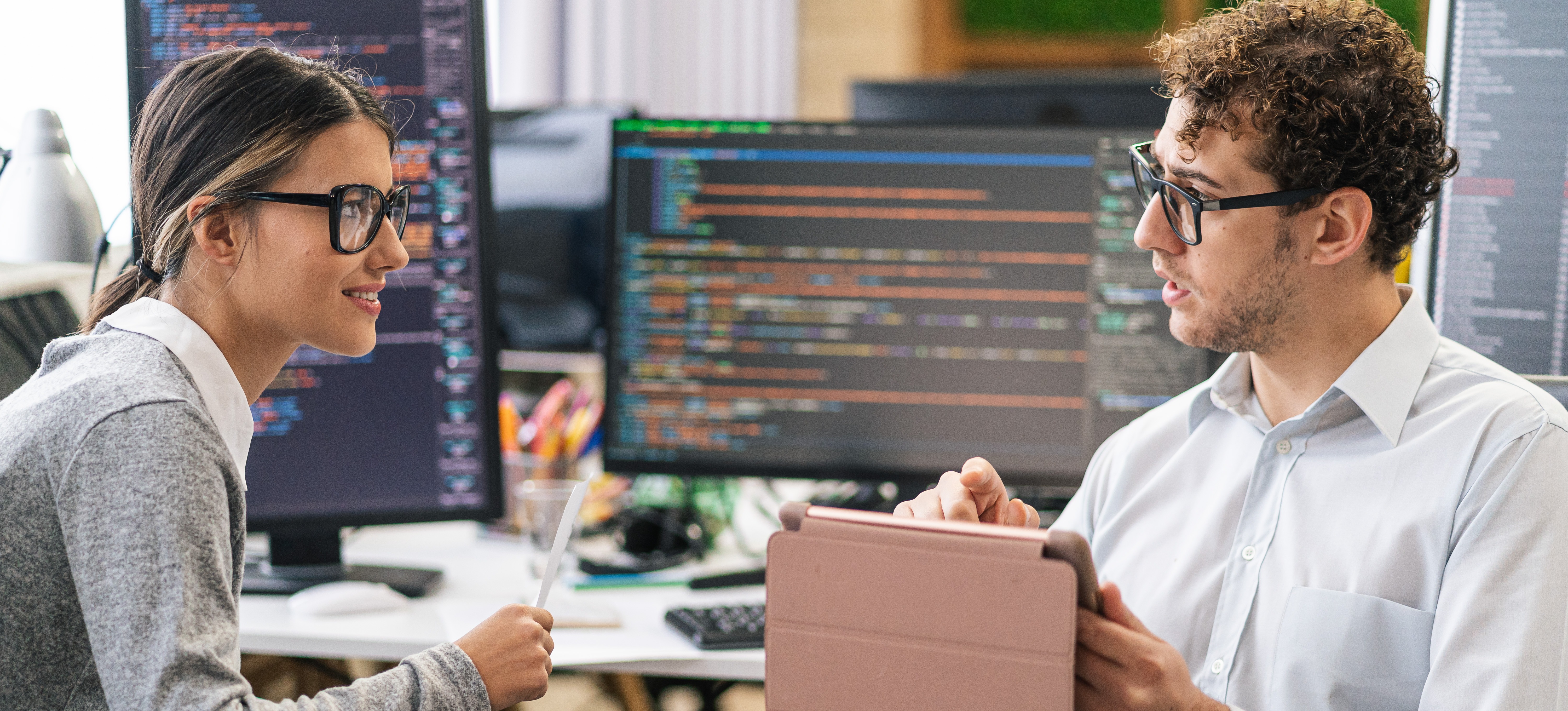 [Hauptbild] Zwei Programmierer diskutieren die Verwendung der Eigenvektorzentralität, während sie in einer Büroumgebung vor mehreren Computermonitoren sitzen.