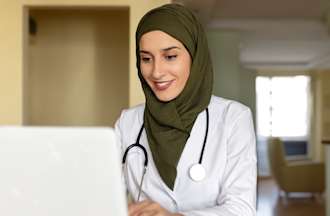 [Featured image] A surgeon in a white coat and stethoscope checks files in front of a computer monitor.