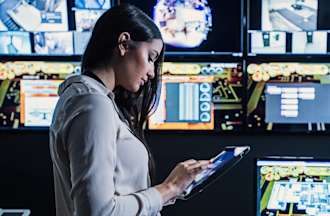 [Featured Image] A person looks at their tablet with computers surrounding them.