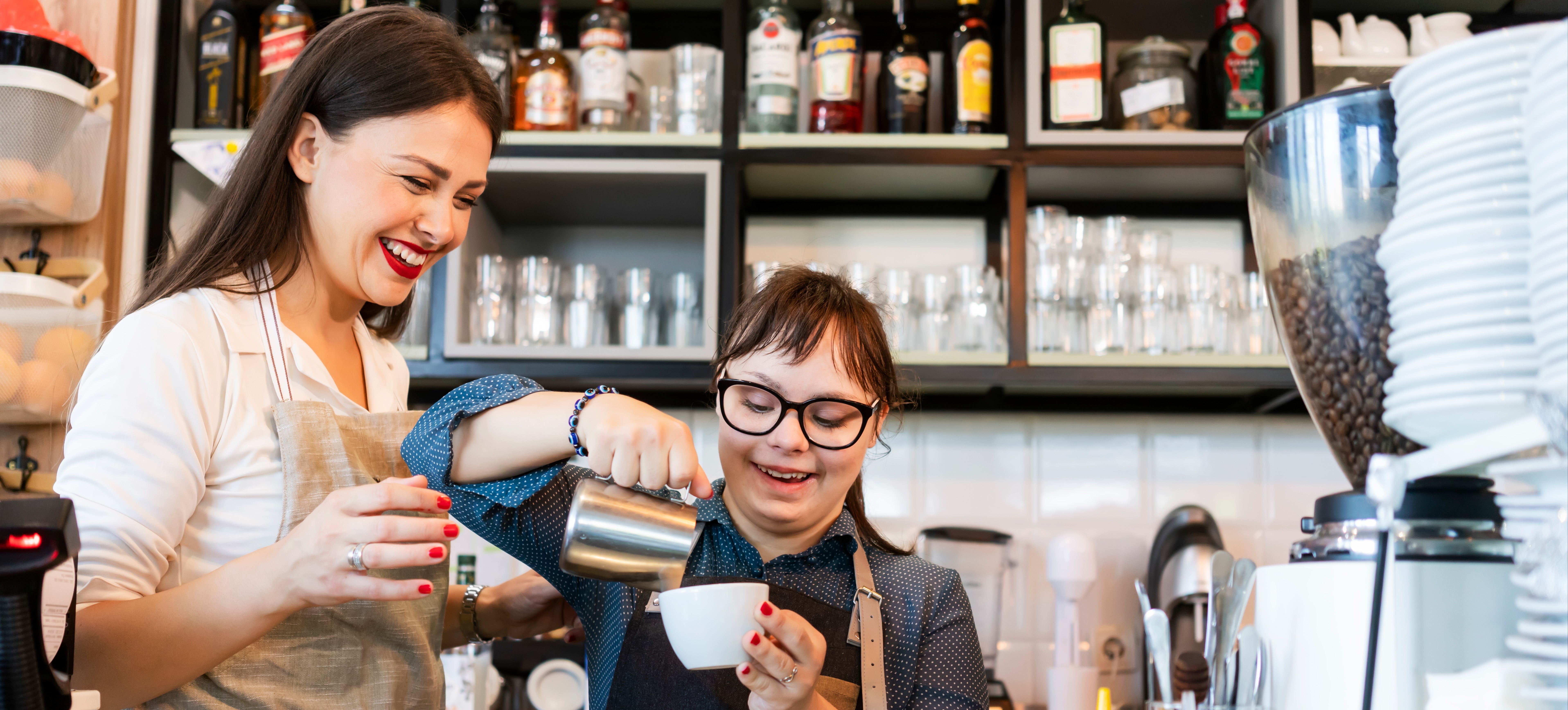 [Featured image] A smiling job coach in an apron stands ready to help a worker with Down Syndrome who is making a cup of coffee in a coffee shop.
