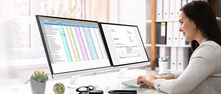 [Featured Image] A smiling person, after discovering how to learn excel, sits at their desktop computer and works on invoices and spreadsheets.
