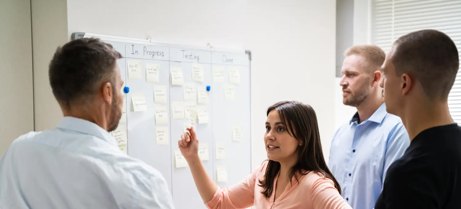[Featured Image] A manager discusses conflict management with their team in front of a whiteboard.   