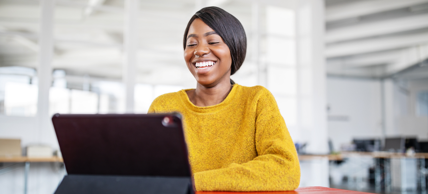 [Featured image] Job candidate smiles at a tablet as they log on for a video interview with a hiring manager in an open office space.