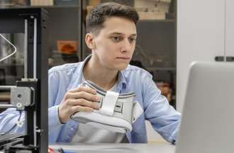 [Feaured image] A young white man looks at his laptop while holding a VR headset. 