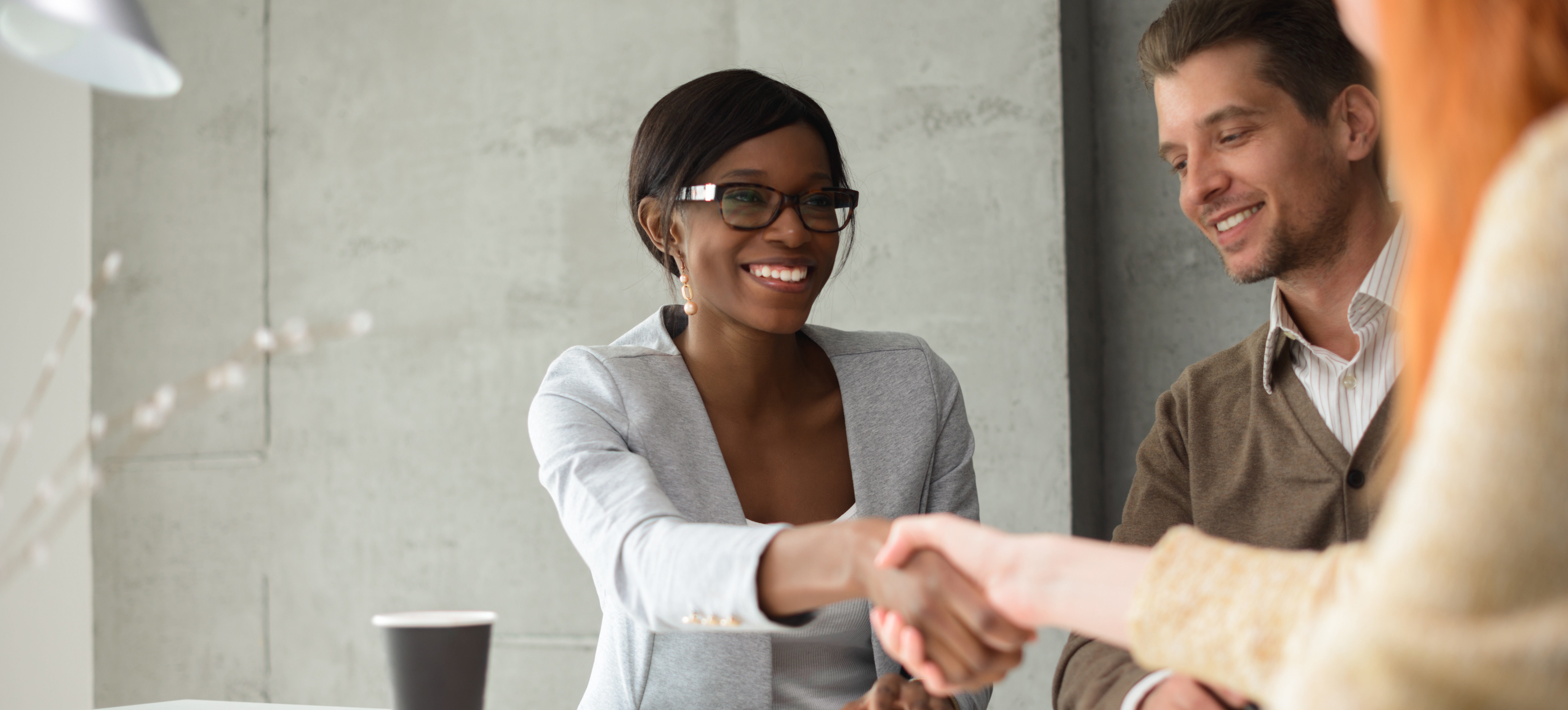 [Featured Image]: Using her sales strategies, a saleswoman shakes the hand of her new client while sitting in an office setting.

