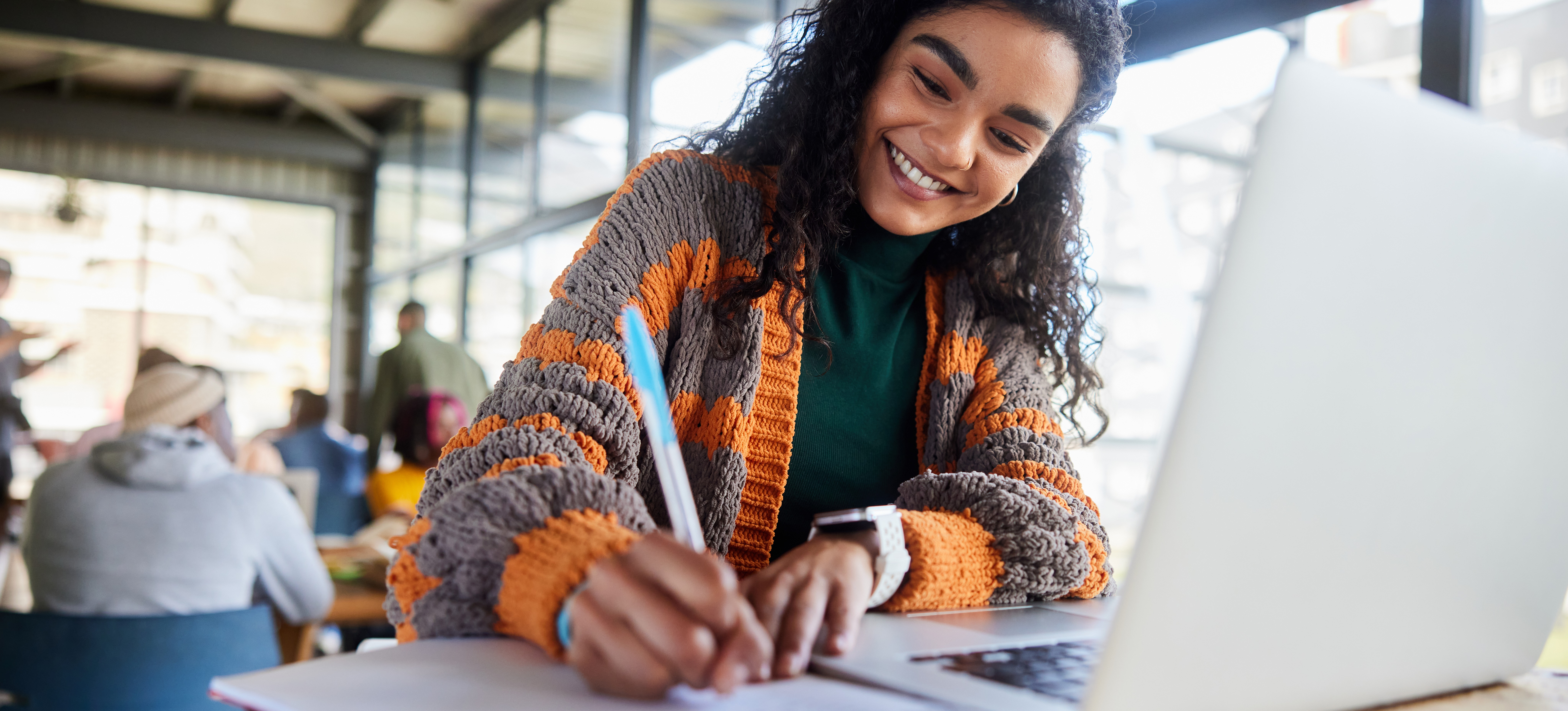 [FEATURED TEXT] A student is sitting at a table with a laptop and notebook in front of them. They are smiling and taking notes in their notebook.
