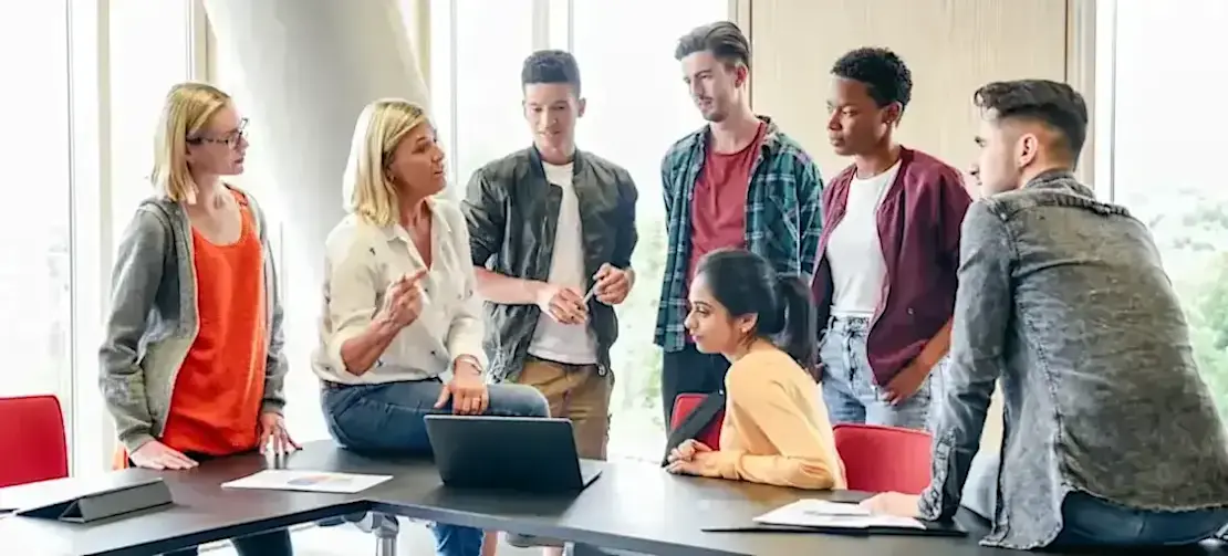 [Featured Image] A group of college students gathers around their professor in a classroom as they explain the relationship between cybersecurity and UX design. 
