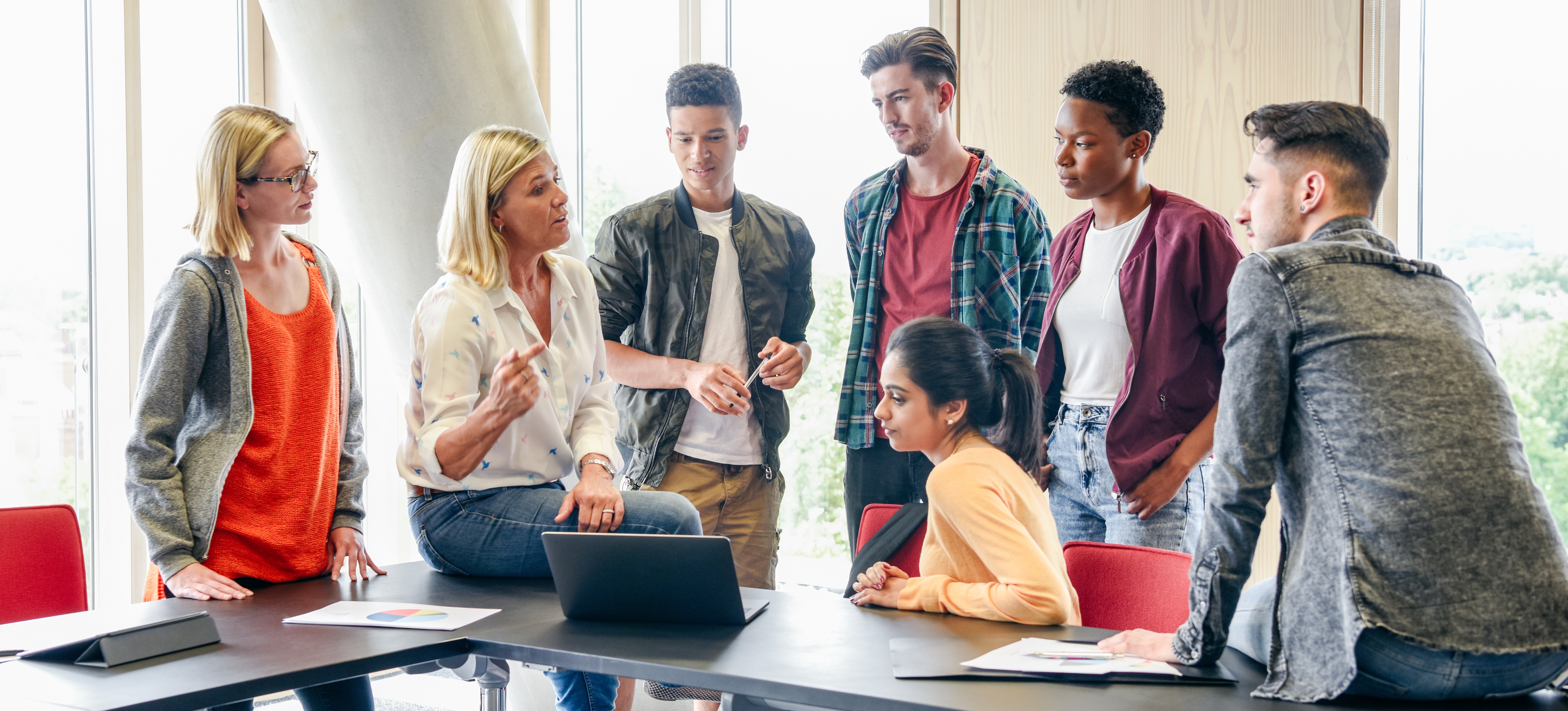 [Featured Image] A group of college students gather around their professor in a classroom as she explains the relationship between cybersecurity and UX design. 
