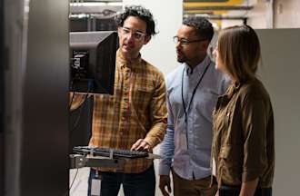 [Featured Image] Three coworkers gather around a desktop computer station in a networking room.