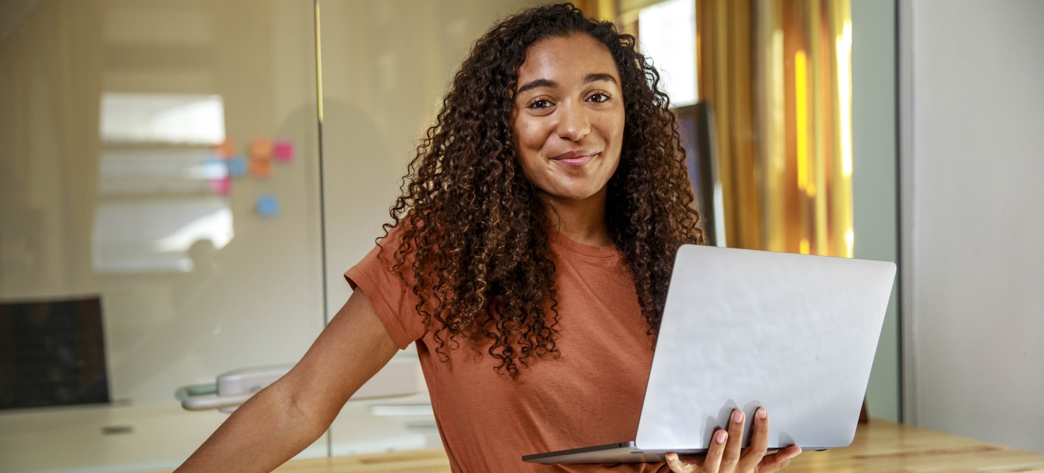 [Featured image] A young brand manager with dark curly hair stands confidently. She holds an open laptop in one hand and leans on the other. 