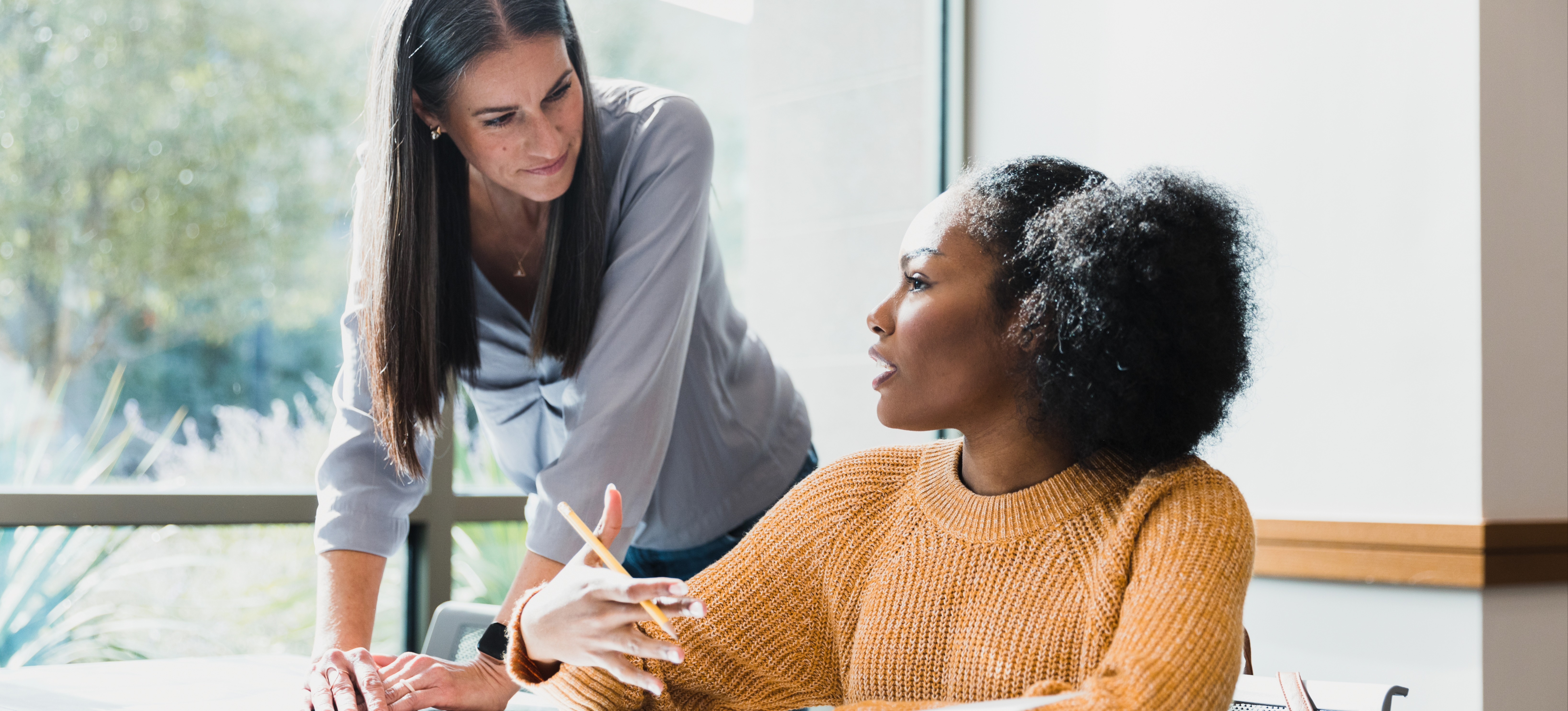 [Featured Image] A college student sits at a table and fills out her college application checklist while consulting a school counselor.

