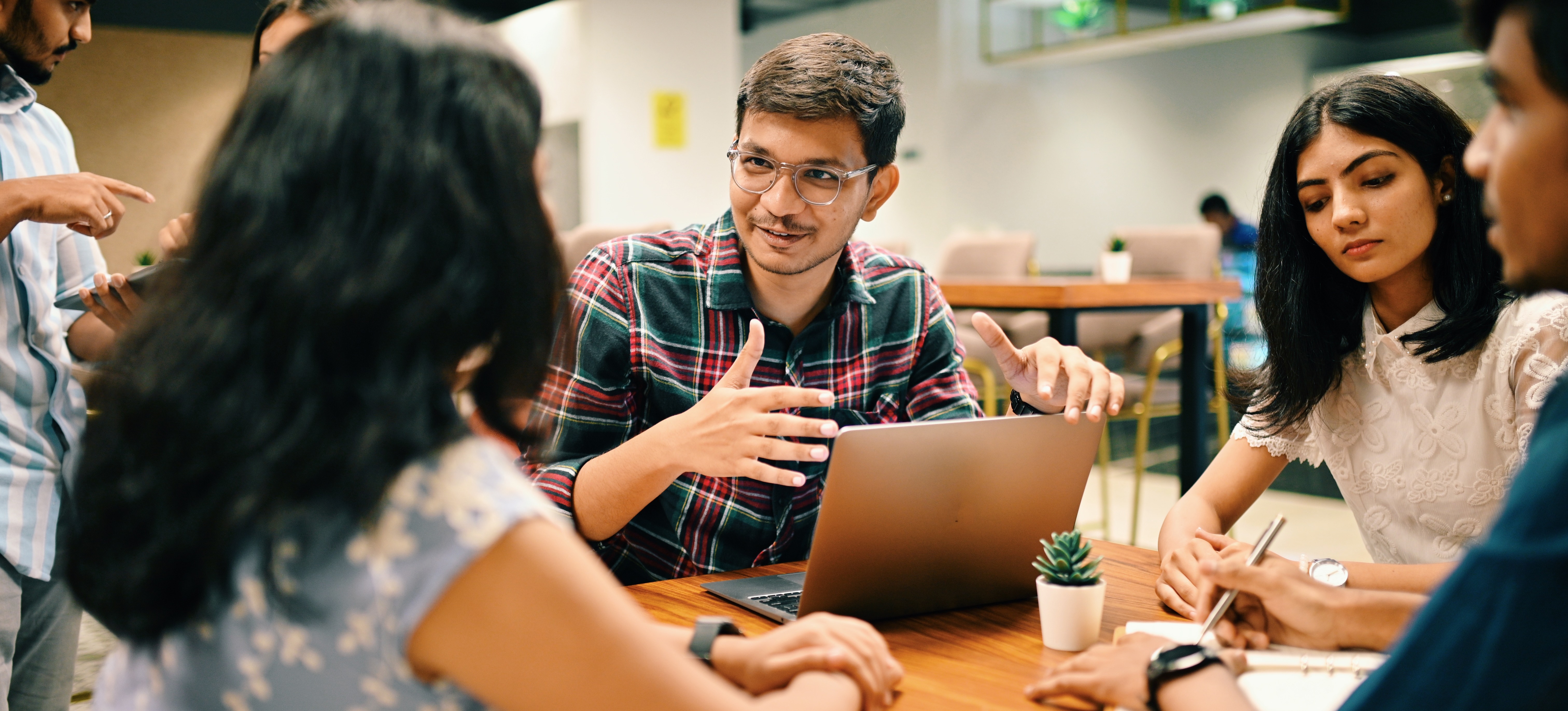 [Featured Image]: A professional in front of a laptop sits around a table with colleagues and practices using effective communication skills.
