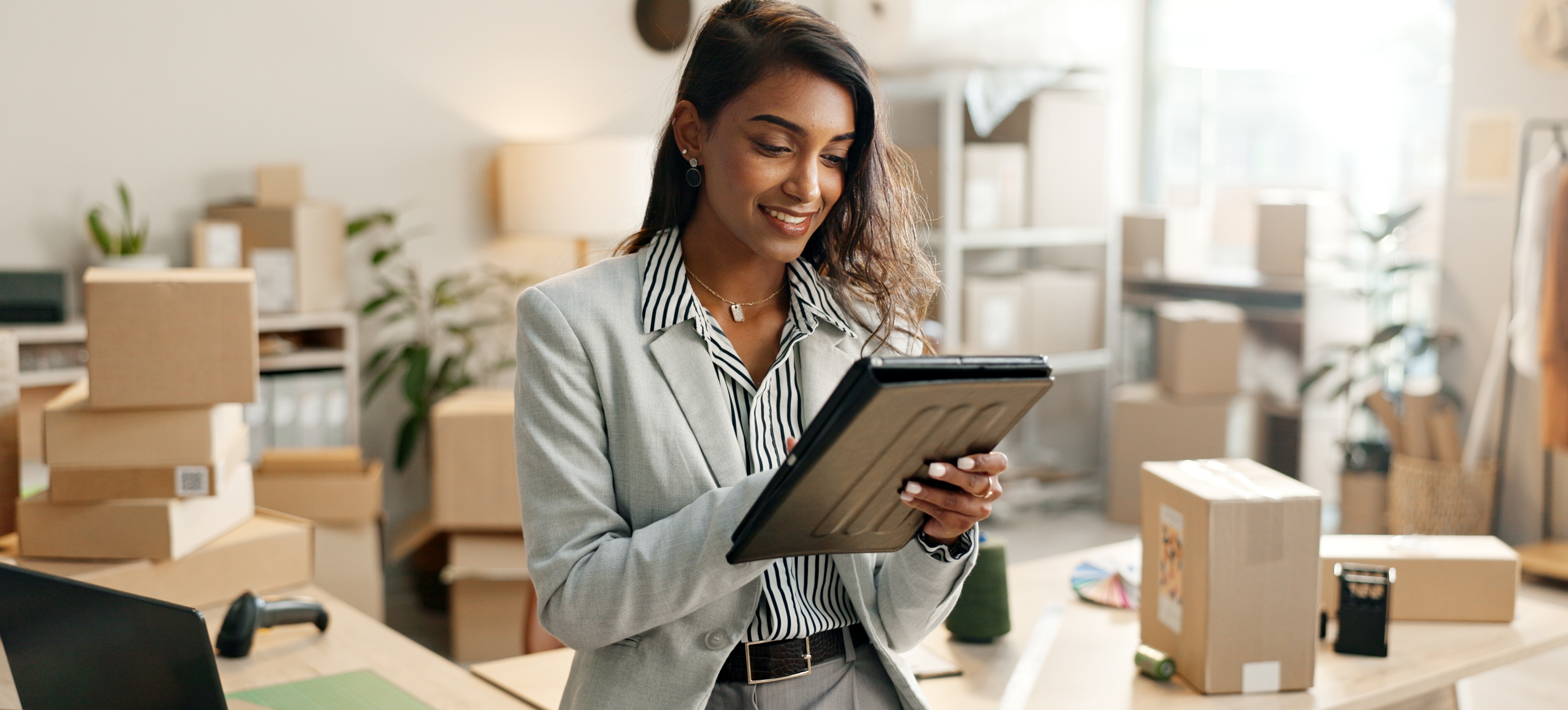[Feature Image] A business owner researches the steps for creating an LLC on their tablet with boxed supplies and equipment in the background. 

