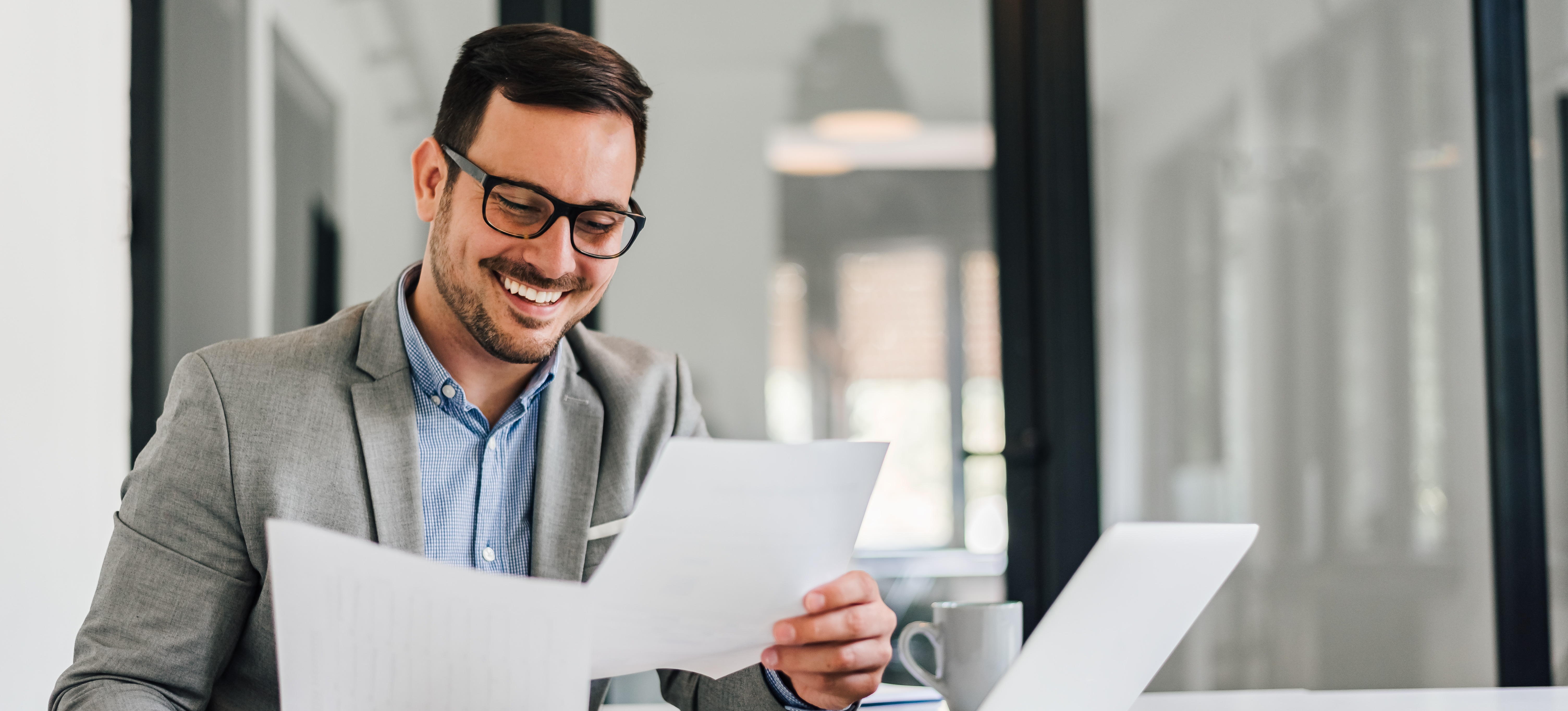 [Featured Image] A cheerful businessman in a suit and sitting in front of his laptop in an office reads an executive summary printed on two sheets of paper.
