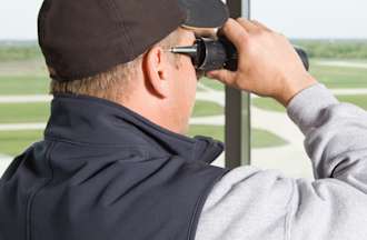 [Featured Image] An air traffic controller, which is one of many jobs that pay $50 an hour without a degree, uses binoculars to look out a window over a runway.
