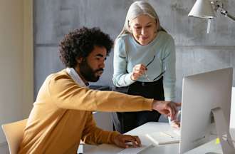 [Featured image] Two coworkers discuss a project at a desktop computer. 