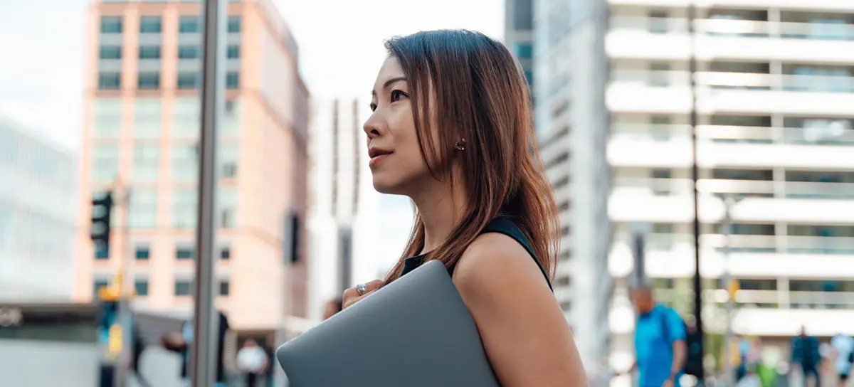 [Featured Image] A professional affected by cyclical unemployment walks on a downtown sidewalk with their laptop in hand on their way to a job interview.
