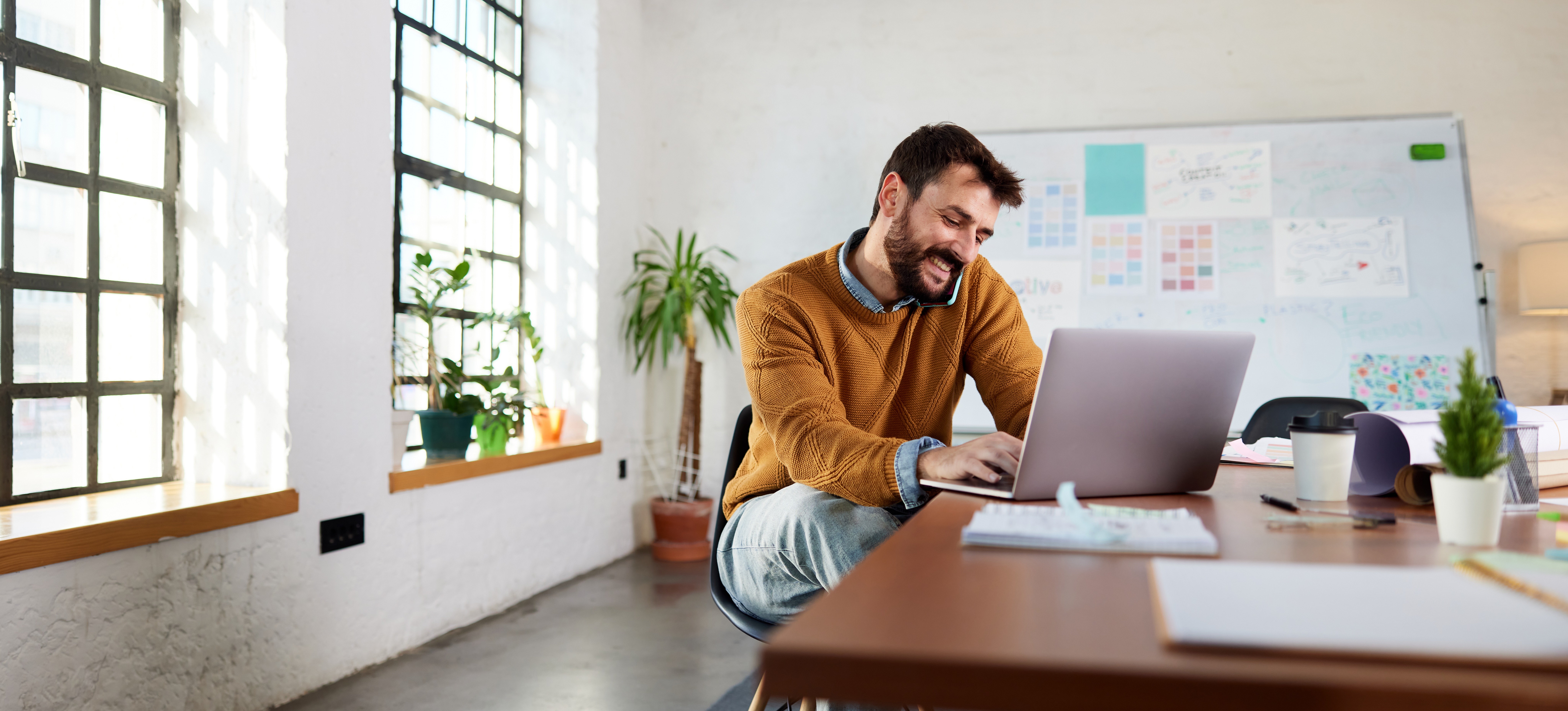 [Feature Image] An interaction designer tests their latest project on a laptop computer while talking on the phone with a colleague in an office with a project board in the background. 
