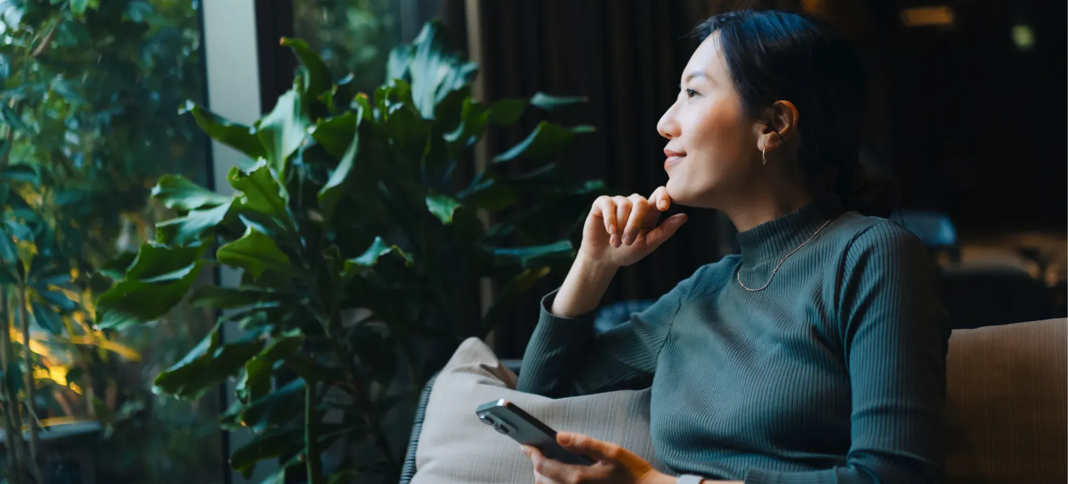 [Featured image] A smiling person in a green sweater sits on a comfortable couch surrounded by indoor plants and looking out a window, contemplating how to use ChatGPT for personal development.
