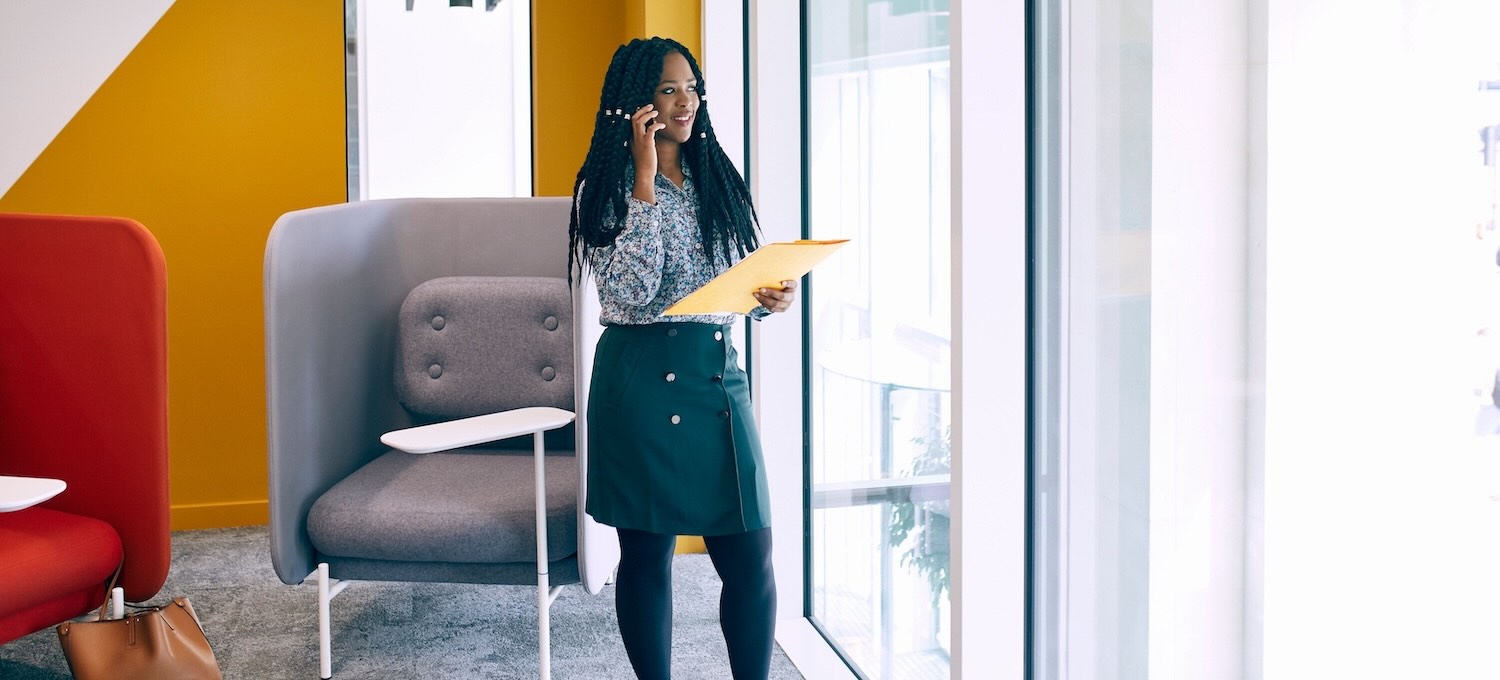 [Featured Image] A web developer who has gotten a job with an associate degree stands in a colorful office, looking out a window, talking on a phone call and holding a yellow folder.