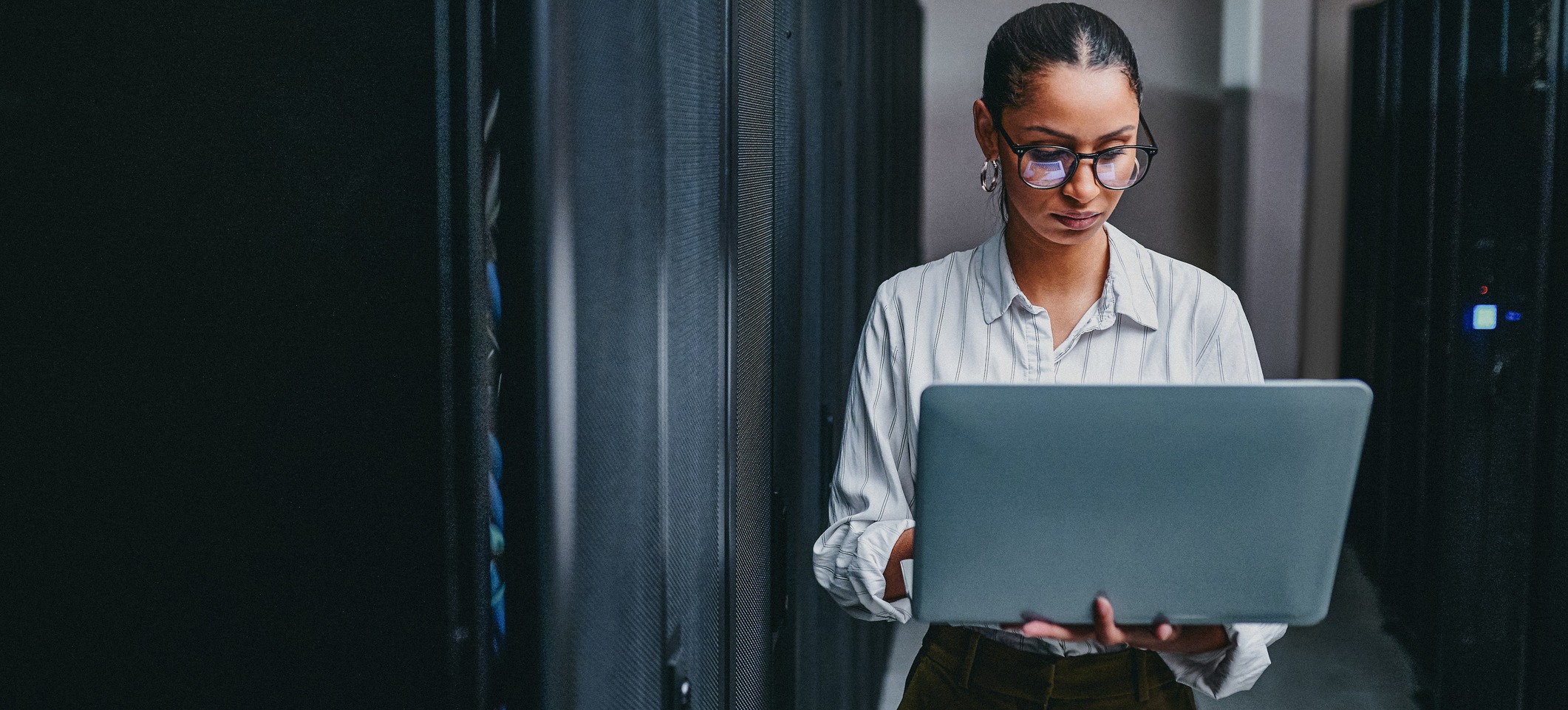  [Hauptbild] Eine IT-Support-Fachkraft mit Brille arbeitet in einem Serverraum an ihrem Laptop.