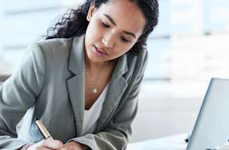 [Featured Image] A pricing analyst takes notes while working at her desk in an office.