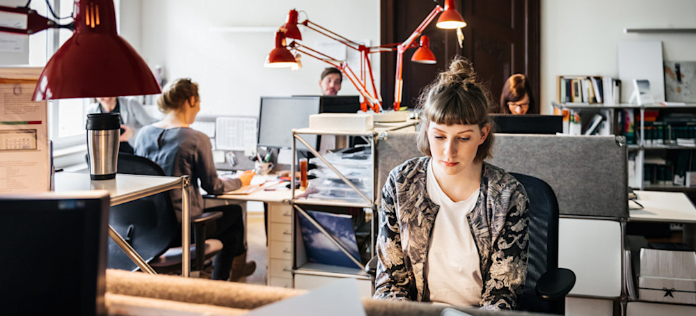[Featured Image] Several start-up software company employees sitting at their desks after a group session applying design thinking to a challenge with the latest iteration of their application. 

