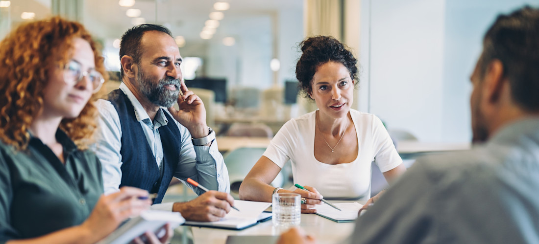 [Featured Image] Four colleagues sit at a conference table in an office setting discussing work matters as the discussion leader uses effective leadership skills to engage the others in listening intently.
