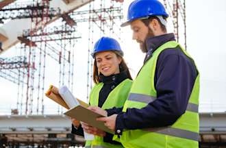 [Featured image] Two engineers in blue hard hats and neon vests work together on a construction site while looking at blueprints.