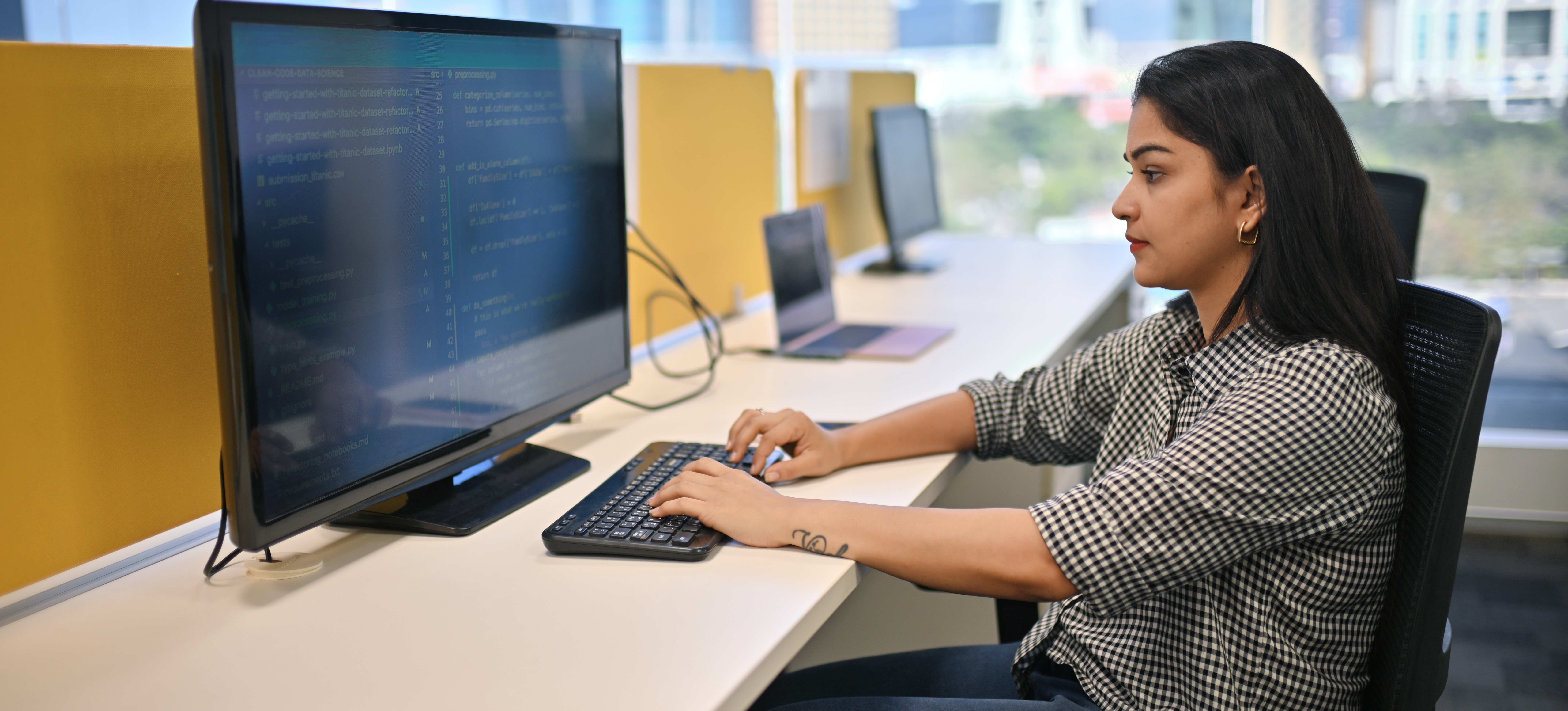 [Featured Image] A blockchain developer uses a computer in an office with large windows showing the cityscape.