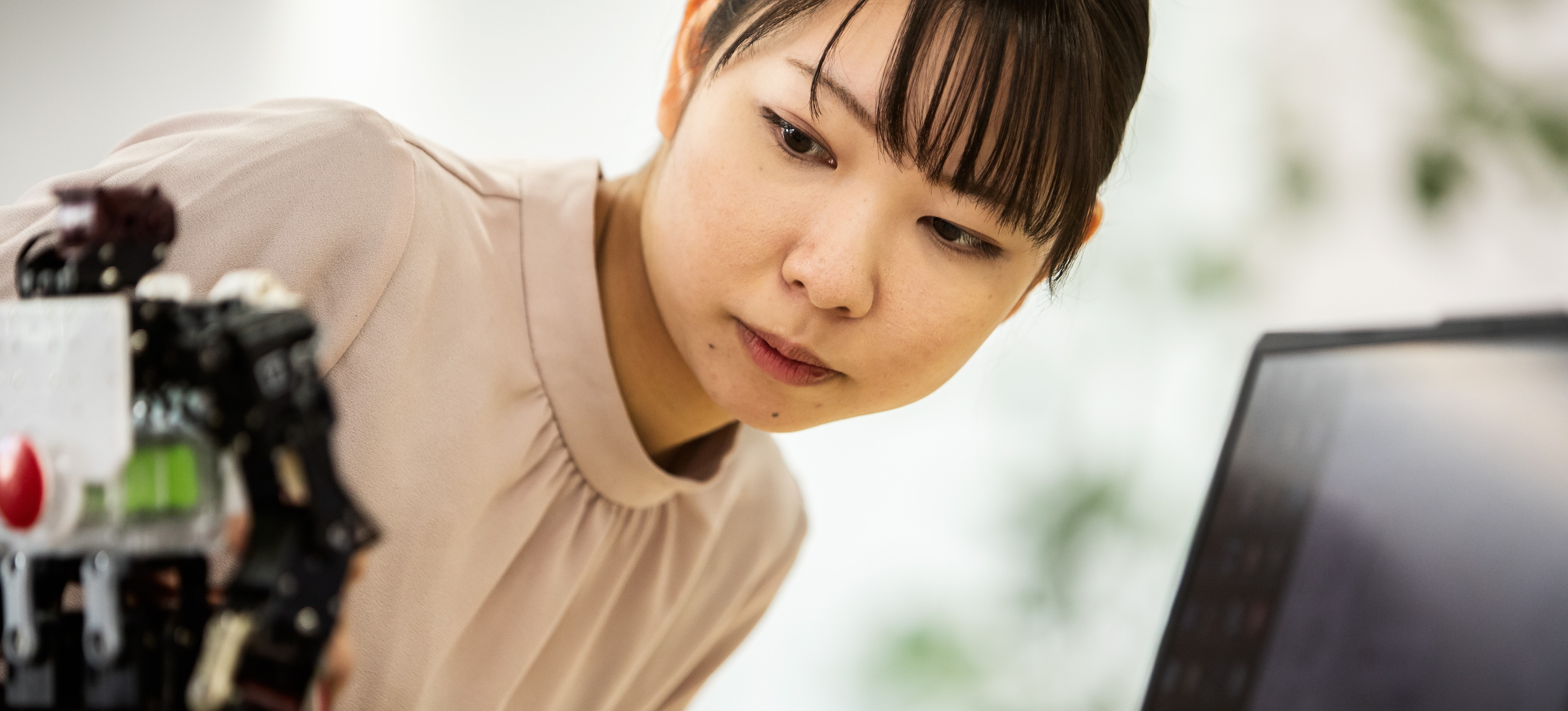 [Featured Image] A woman uses reinforcement learning to fine-tune her robotics work as she examines a small robot next to an open laptop.
