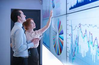 [Featured image] Two MBA finance students examine financial graphs on a backlit whiteboard