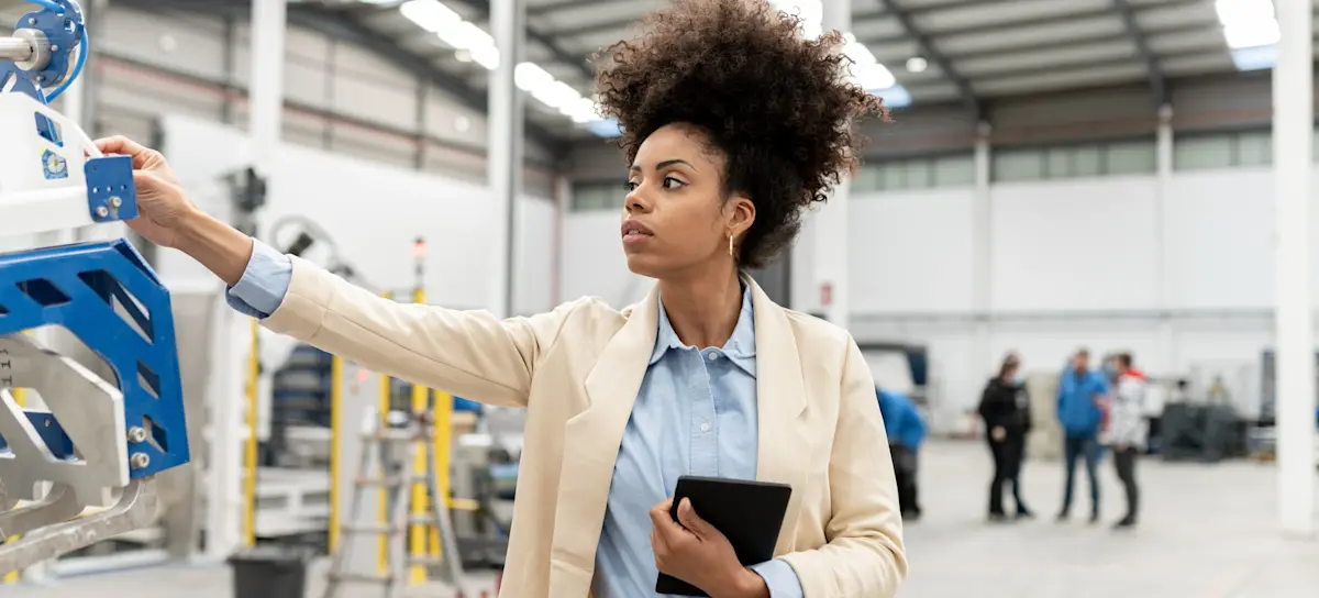 [Featured Image] A person working for an automation engineer salary examines a piece of machinery in a factory setting while holding a tablet.
