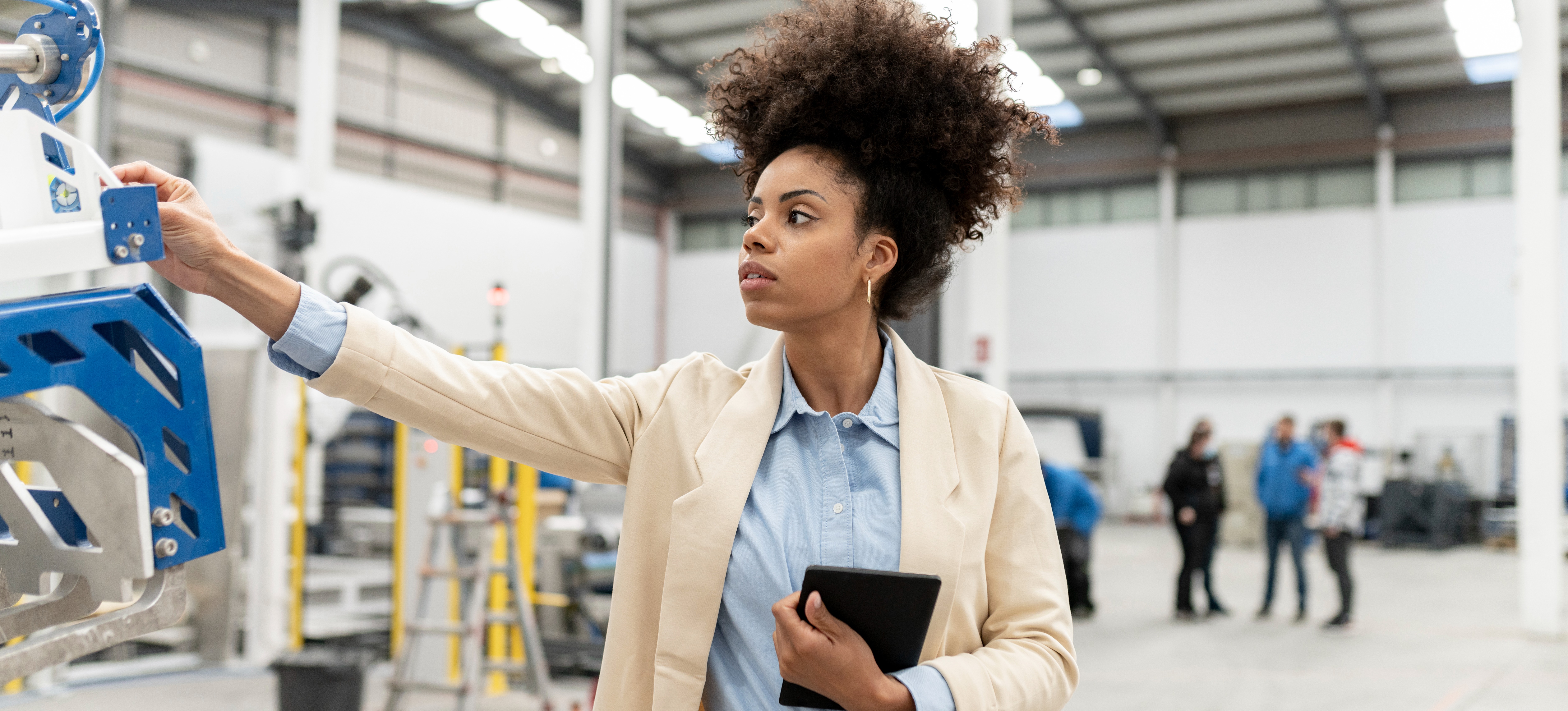 [Featured Image] A woman working for an automation engineer salary examines a piece of machinery in a factory setting while holding a tablet.
