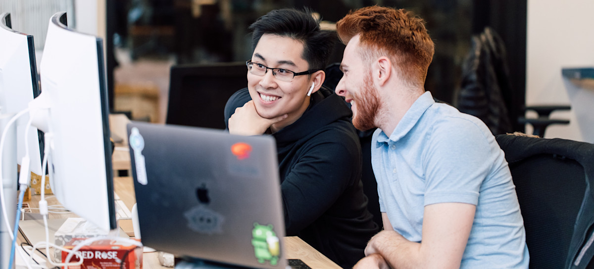[Featured image] Two video game designers are working together on a laptop and desktop in an office.