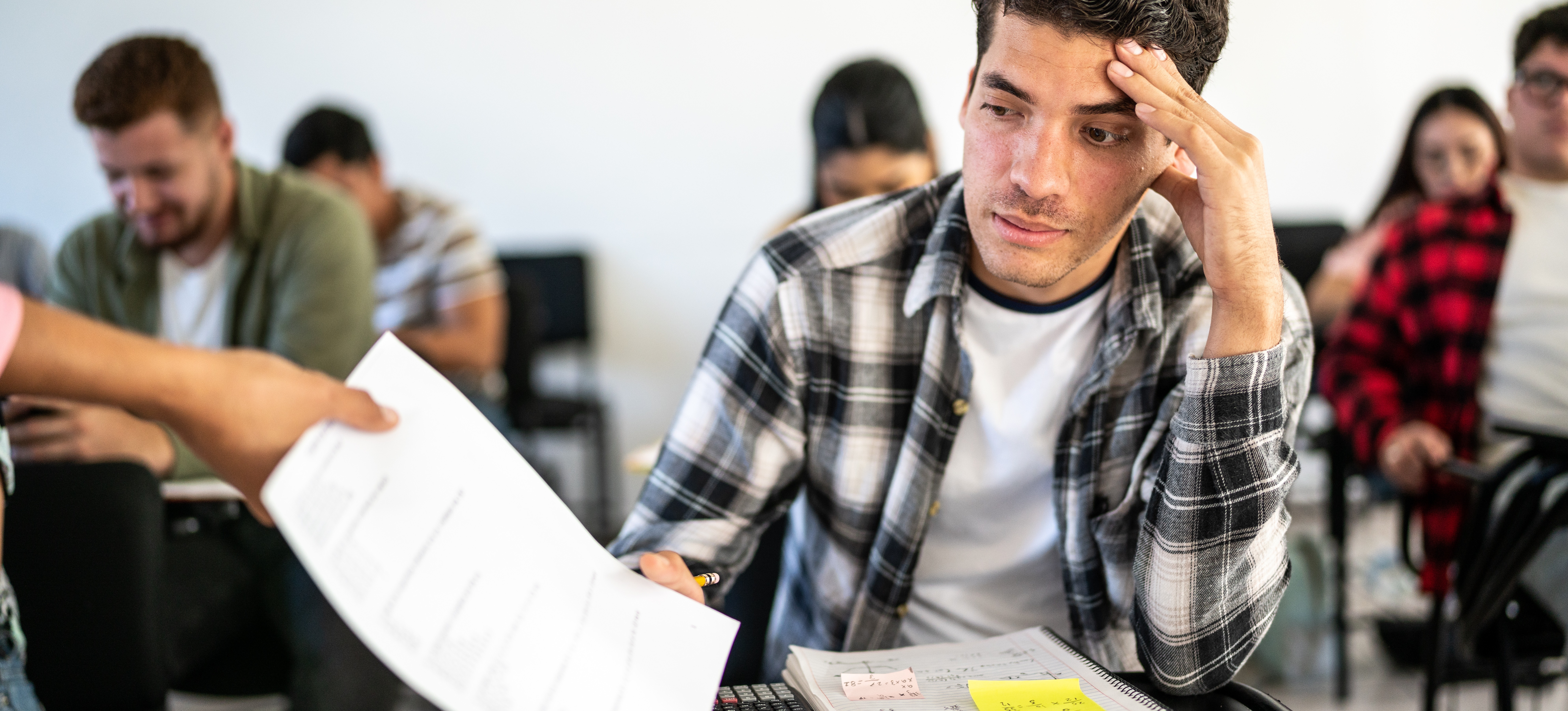 [Featured Image] A male college student facing academic failure disappointedly receives a graded paper from a professor during class.